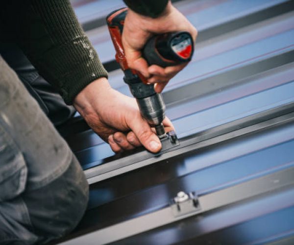 Building contractor is installing metal roofing sheets on the rooftop of the house using electric screwdriver.