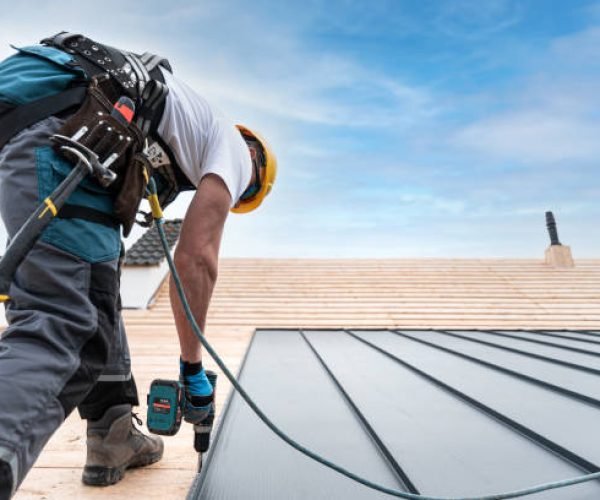 A roofer with a safety harness and tool belt is working with a electric screwdriver on the roof. He is anchoring the metal roofing with a screws.