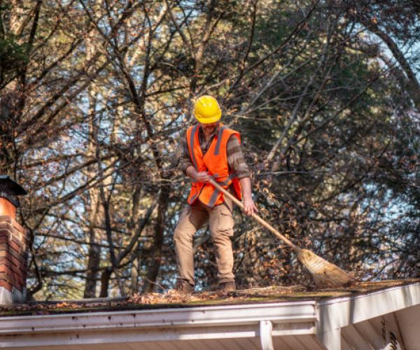 A 53-year-old worker in high visibility orange vest and yellow hard hat performs maintenance on a residential roof. He sweeps fallen autumn leaves while working near a brick chimney, surrounded by bare tree branches against clear blue sky